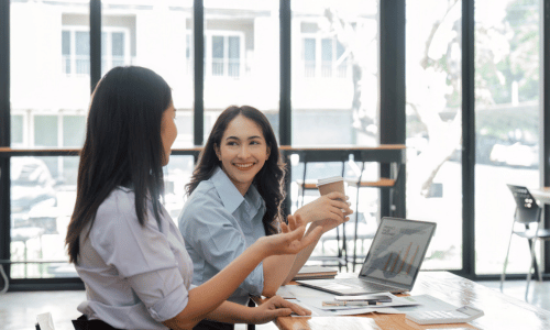 Two colleagues chatting and smiling at a desk with a laptop