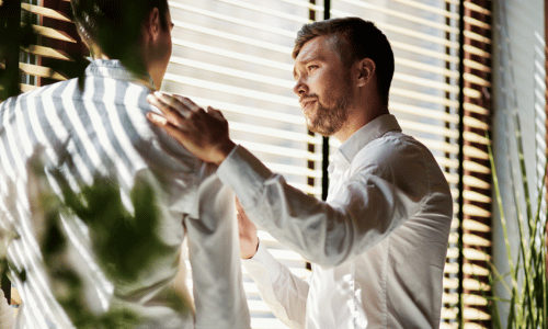A man offering support to a colleague, with a hand on his shoulder, near a sunlit window.